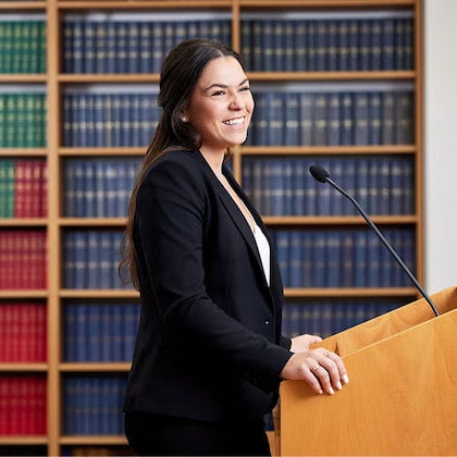 a middle-aged woman in a black and white suit, standing behind a lectern, smiling.