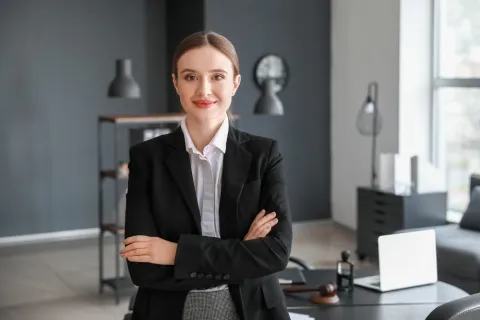 Professional woman in a dark blazer with arms crossed, smiling at the camera in a modern office, with a desk, laptop, and decorative items in the background.