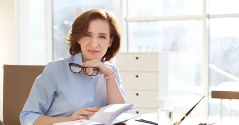 1. A woman wearing glasses sits at a desk, writing in a notebook, focused on her work.