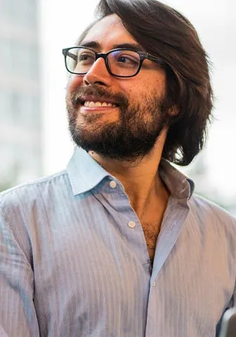 A man with glasses and a beard stands in front of a window, looking thoughtfully outside.