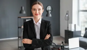 Professional woman in a dark blazer with arms crossed, smiling at the camera in a modern office, with a desk, laptop, and decorative items in the background.