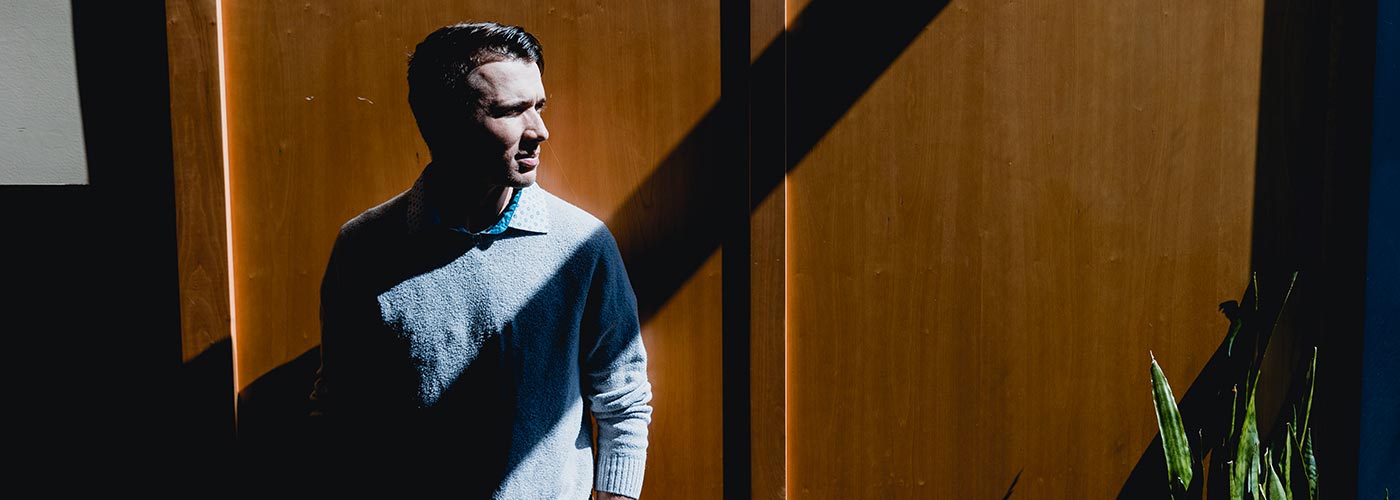 A man stands in front of a wooden door, looking directly at the camera with a neutral expression.